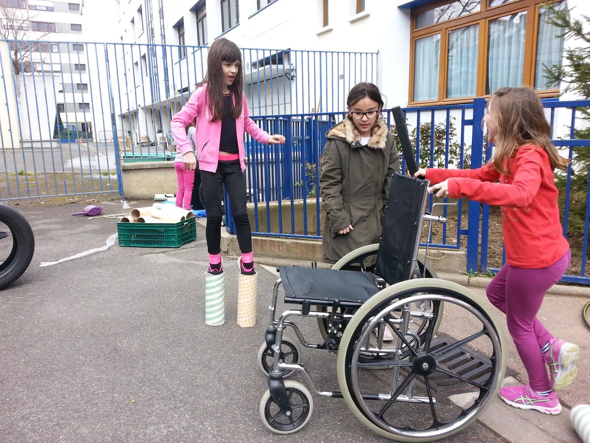 Des enfants jouent avec des échasses et un fauteuil roulant dans la cour